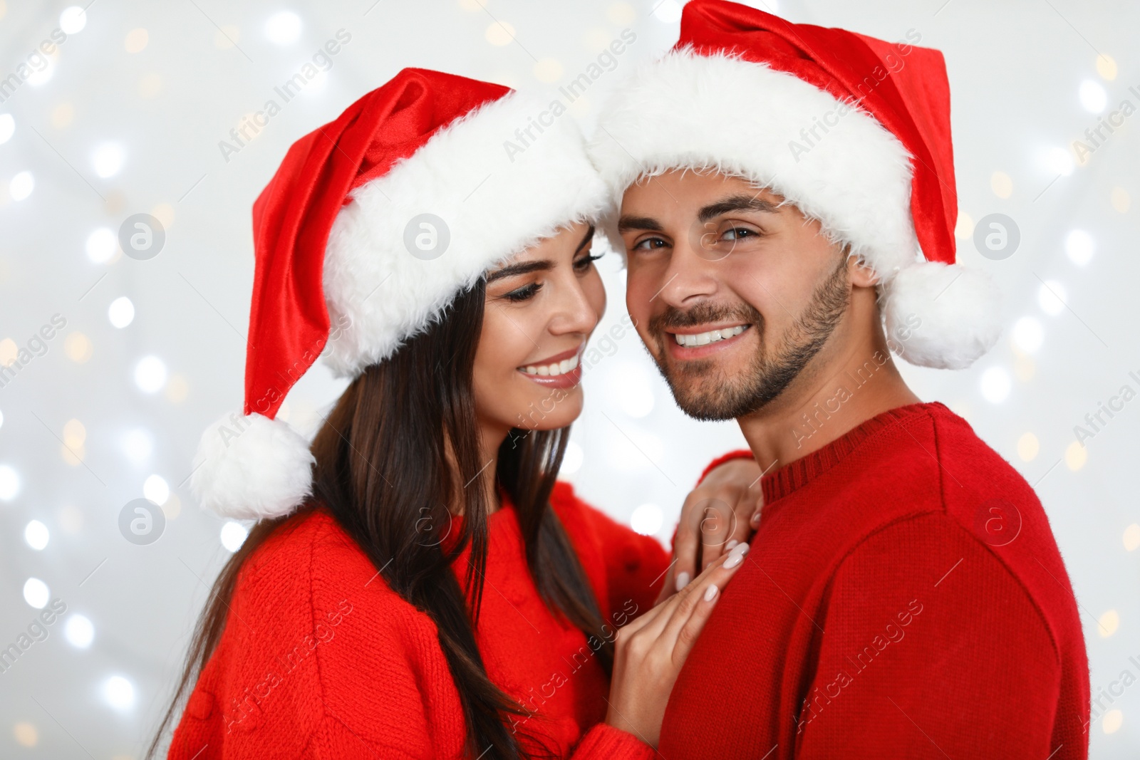 Photo of Lovely young couple in Santa hats against blurred festive lights. Christmas celebration