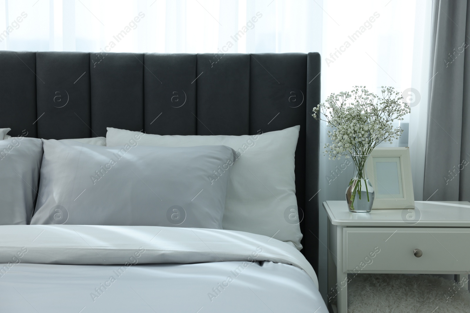 Photo of Vase with beautiful flowers on white nightstand and comfortable bed in room. Stylish interior