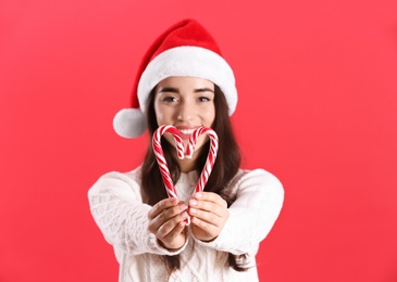Beautiful woman in Santa Claus hat making heart with candy canes on red background