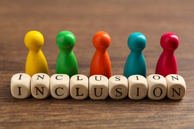 Photo of Colorful pawns and cubes with word Inclusion on wooden table, closeup