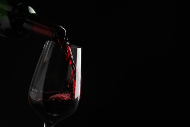 Photo of Pouring tasty red wine from bottle into glass on black background, closeup. Space for text