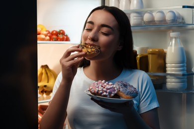 Photo of Young woman eating donut near refrigerator in kitchen at night. Bad habit