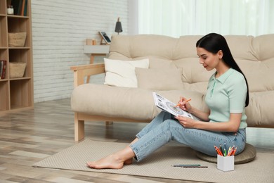 Photo of Young woman coloring antistress page near sofa in living room