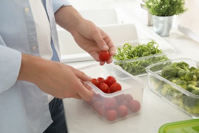 Man putting cherry tomato into plastic container with fresh vegetables in kitchen, closeup. Food storage