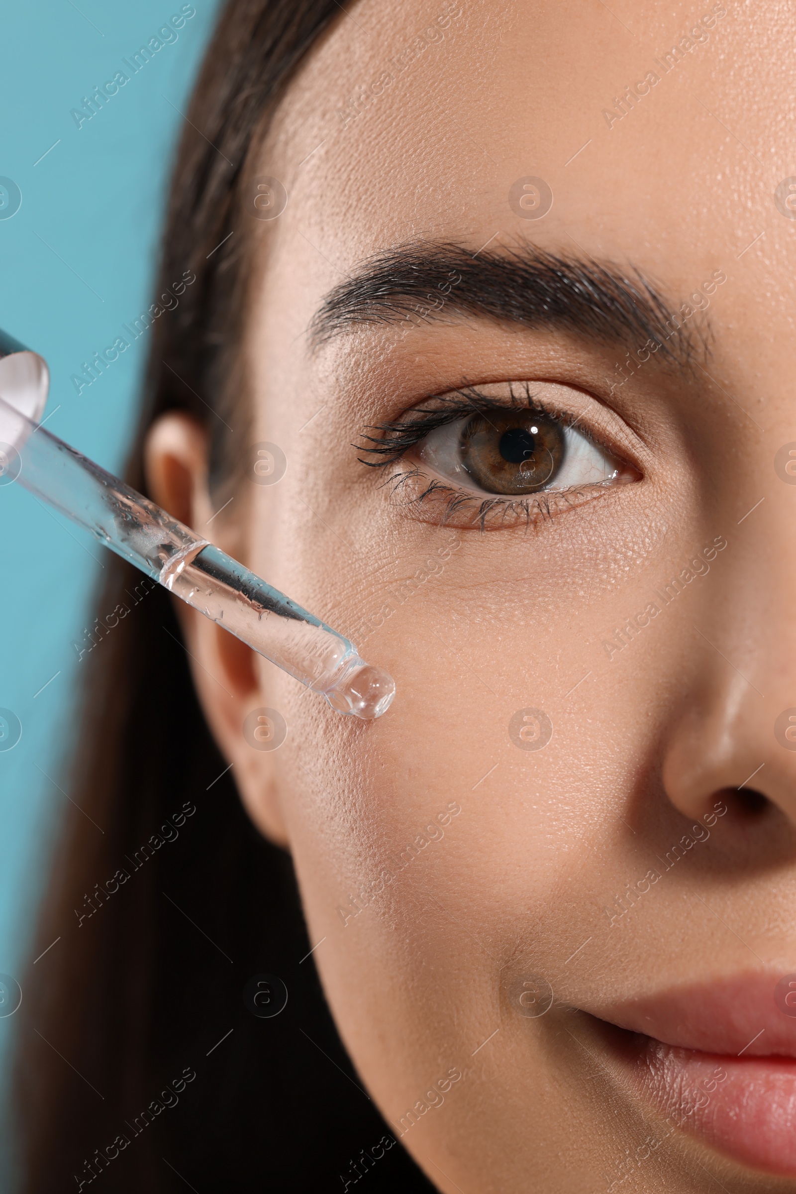 Photo of Beautiful young woman applying serum onto her face on light blue background, closeup