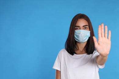 Photo of Young woman in protective mask showing stop gesture on light blue background, space for text. Prevent spreading of coronavirus