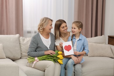 Photo of Little girl congratulating her mom and granny with flowers and postcard at home. Happy Mother's Day