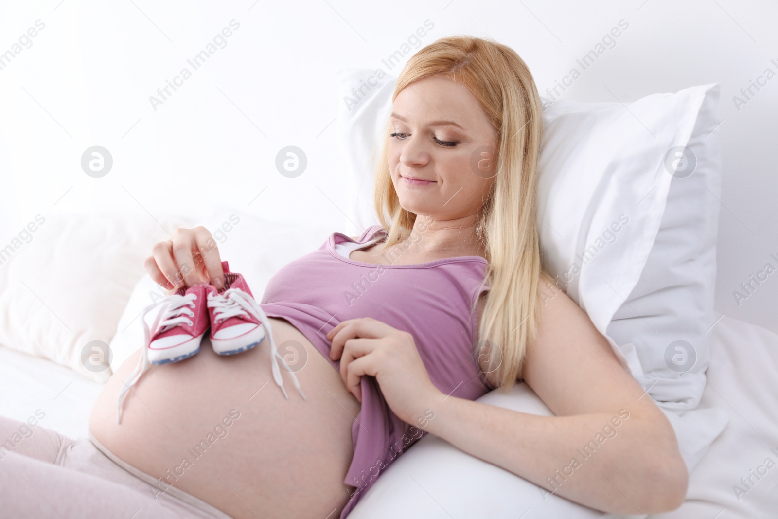 Photo of Beautiful pregnant woman holding baby booties near tummy on bed in light room