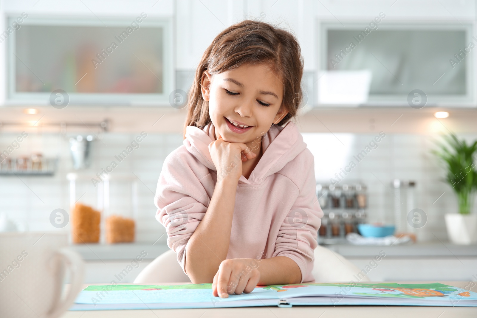 Photo of Cute little girl reading book at table in kitchen