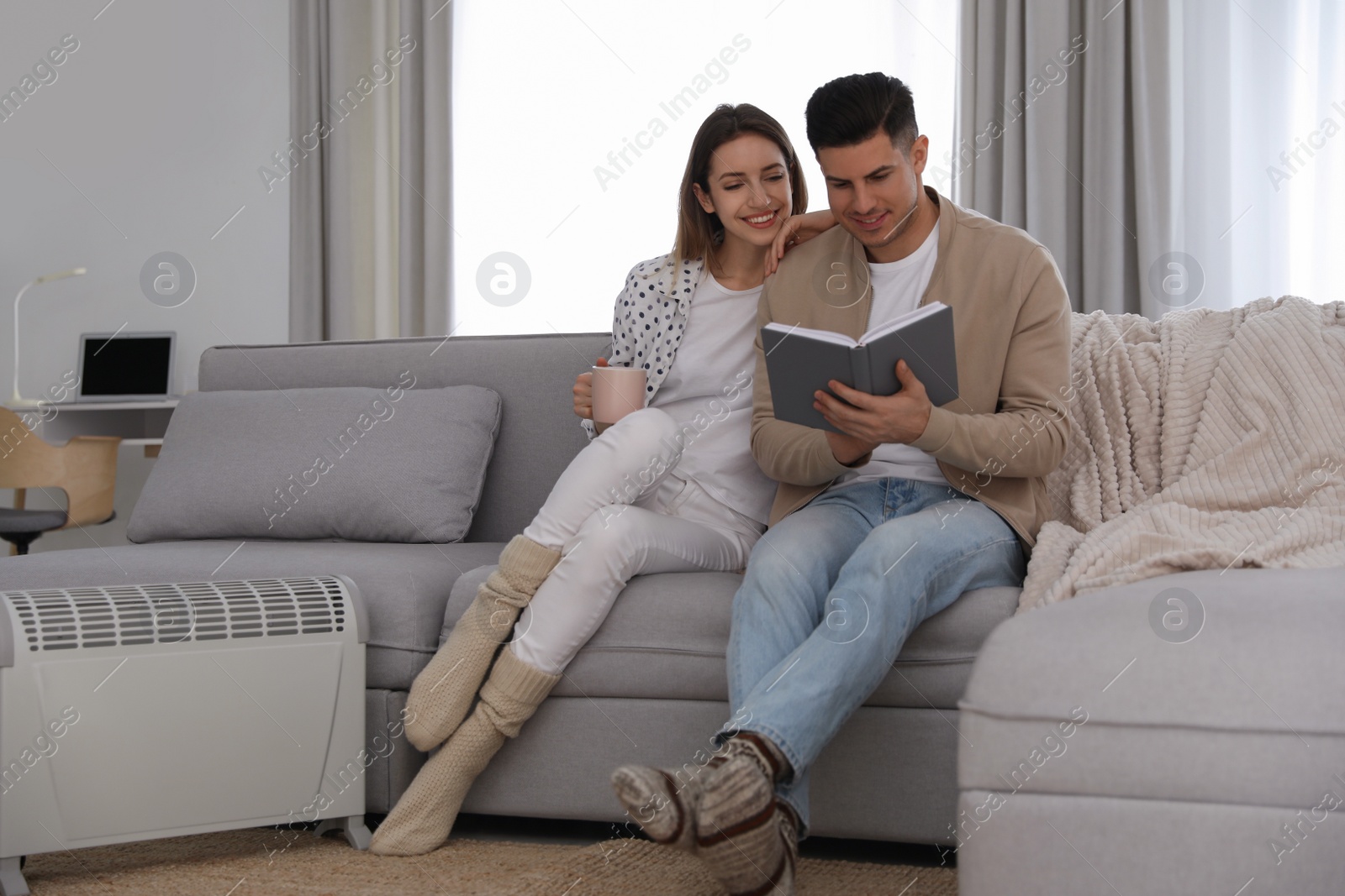 Photo of Happy couple sitting on sofa near electric heater at home