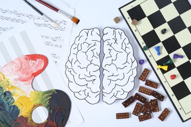 Photo of Logic and creativity. Paper brain on white background, flat lay. Paint palette and music notes near left hemisphere and board games near right one
