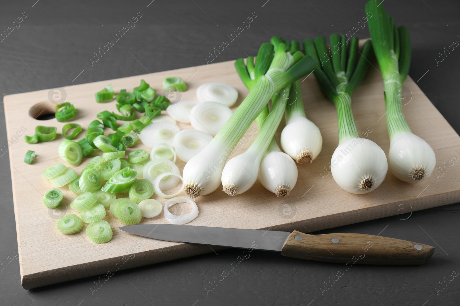 Photo of Board with knife, whole and cut spring onions on grey wooden table, closeup