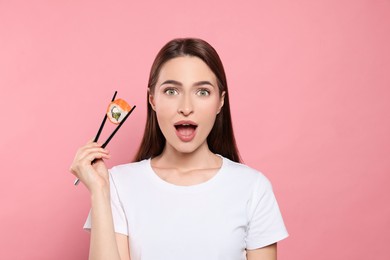 Photo of Emotional young woman holding sushi roll with chopsticks on pink background