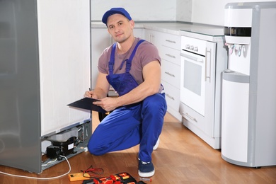 Photo of Male technician with clipboard and tools near broken refrigerator in kitchen