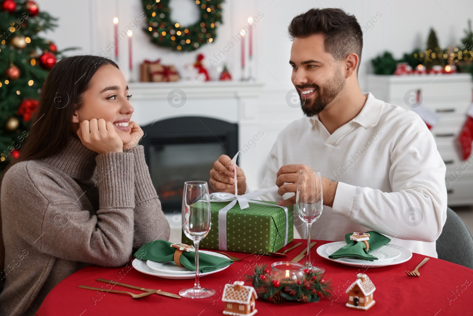 Photo of Happy young man opening Christmas gift from his girlfriend at table indoors
