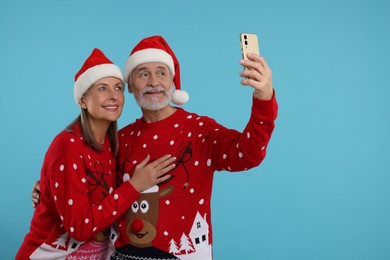 Senior couple in Christmas sweaters and Santa hats taking selfie on light blue background. Space for text