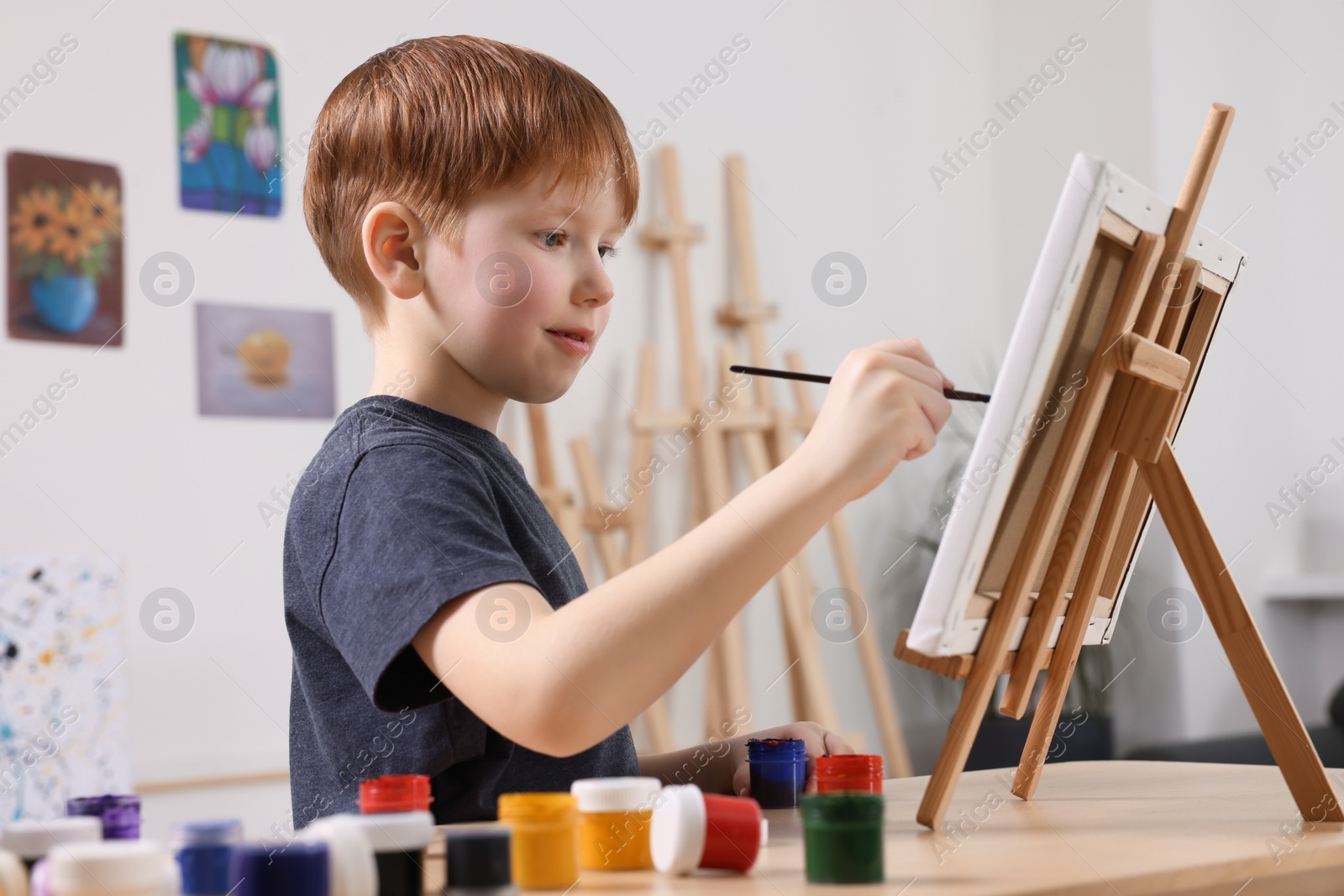 Photo of Little boy painting at table in studio. Using easel to hold canvas