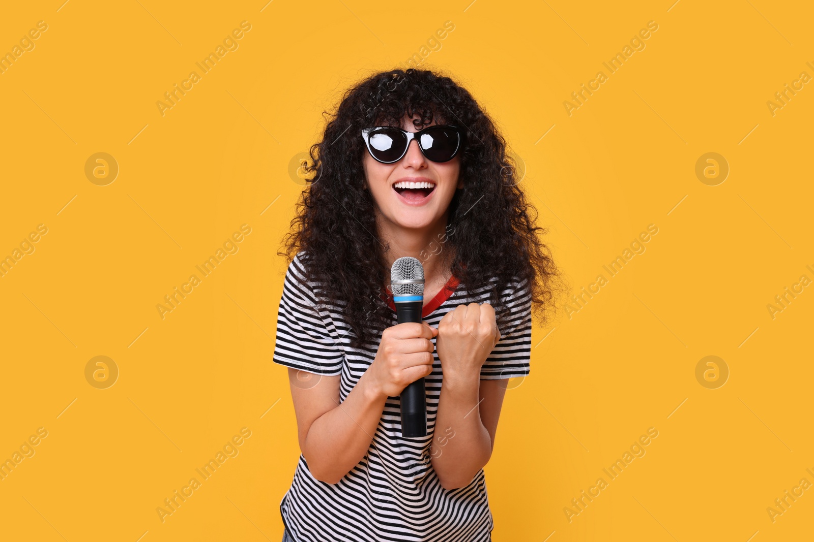 Photo of Beautiful young woman with microphone and sunglasses singing on yellow background