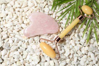 Photo of Quartz gua sha tool, face roller and green leaves on white stones, flat lay