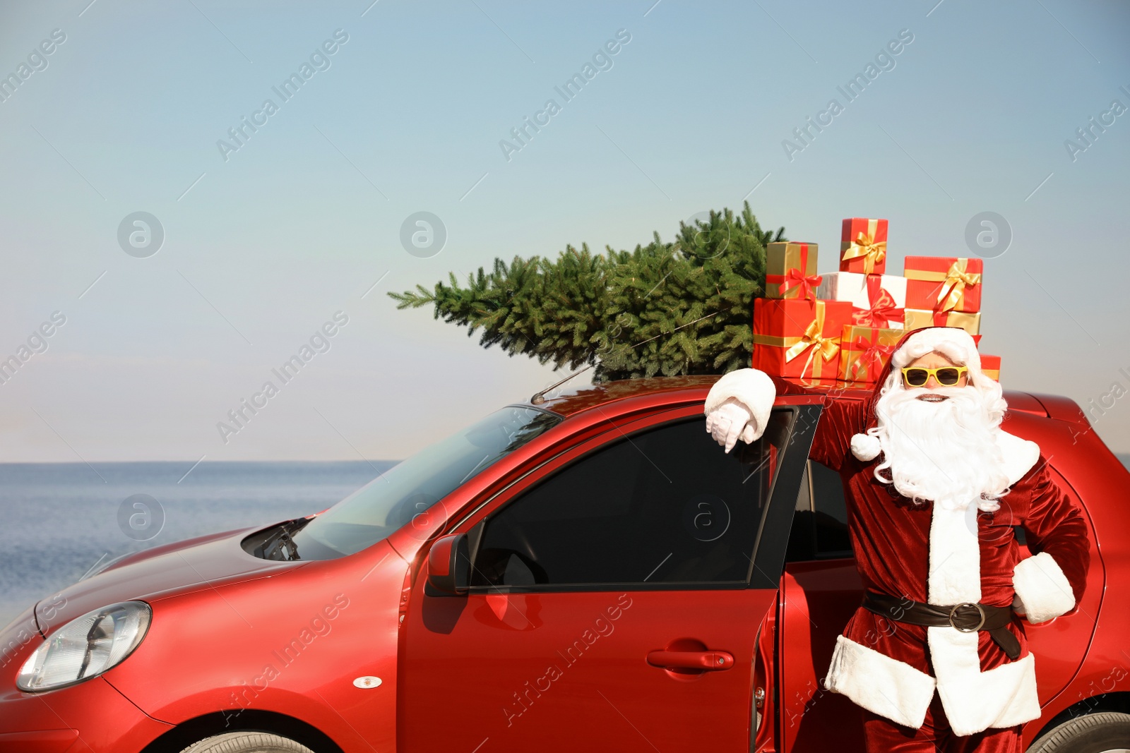 Photo of Authentic Santa Claus near car with presents and fir tree on roof at sea