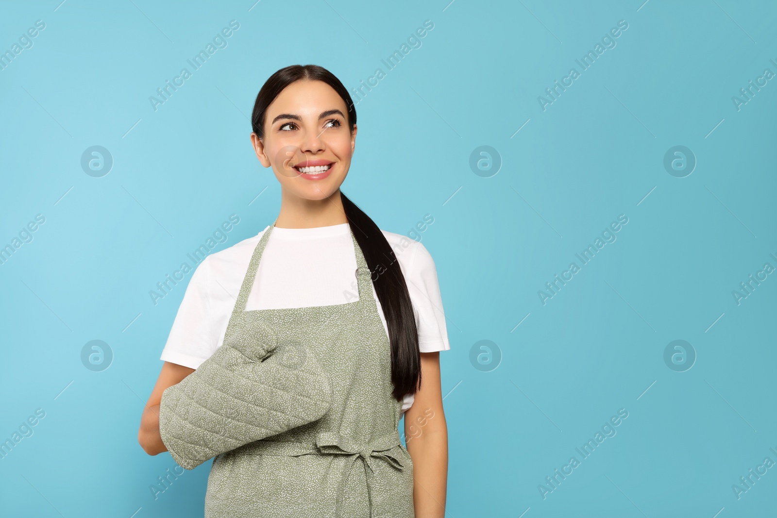 Photo of Young woman in grey apron and oven glove on light blue background, space for text