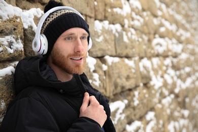 Photo of Young man listening to music with headphones near stone wall. Space for text