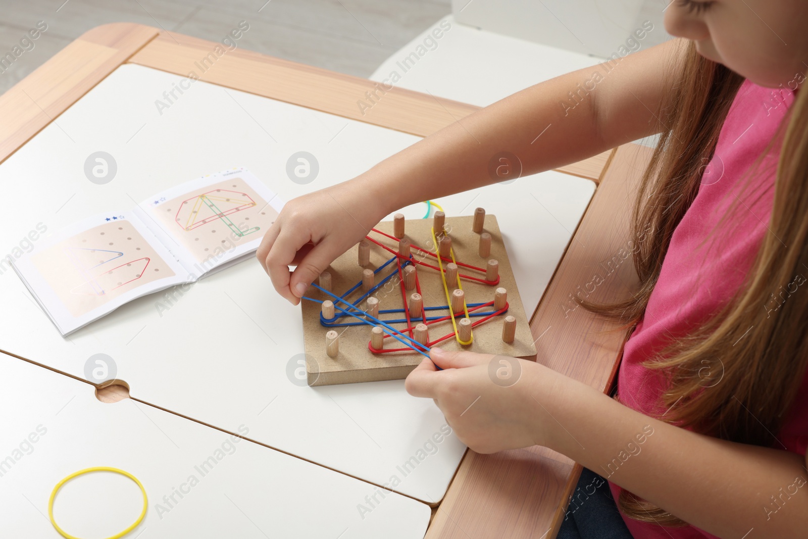 Photo of Motor skills development. Girl playing with geoboard and rubber bands at white table indoors, closeup