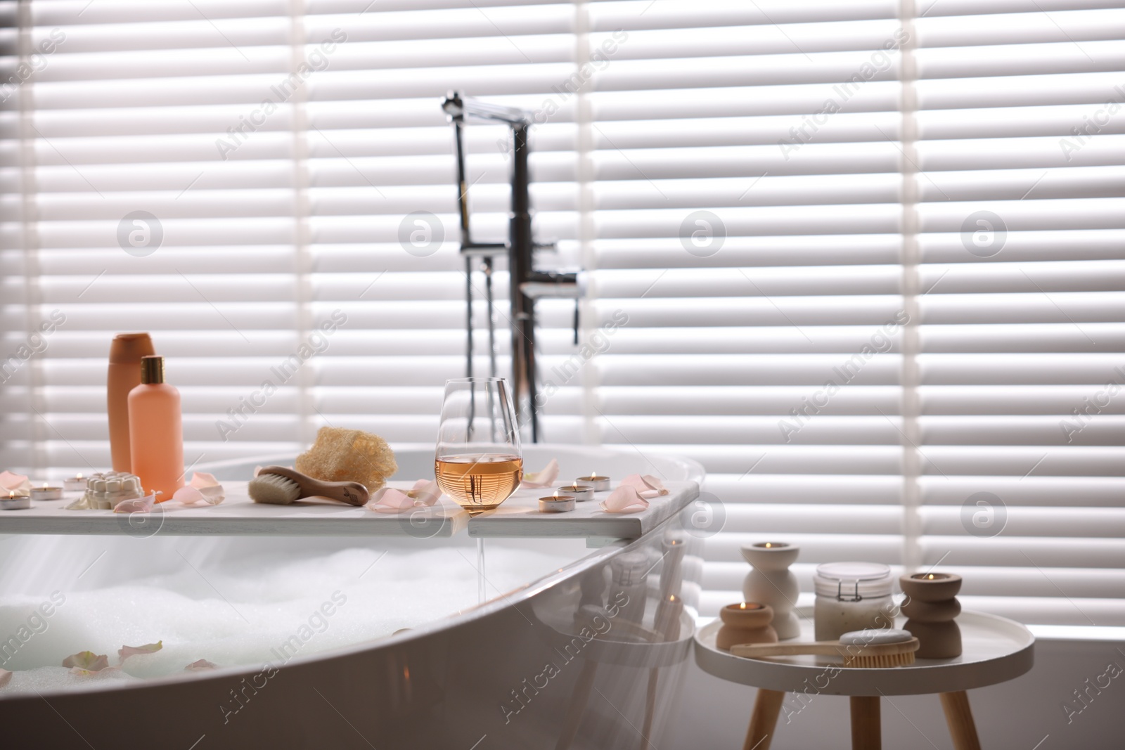 Photo of Wooden tray with wine, toiletries and flower petals on bathtub in bathroom