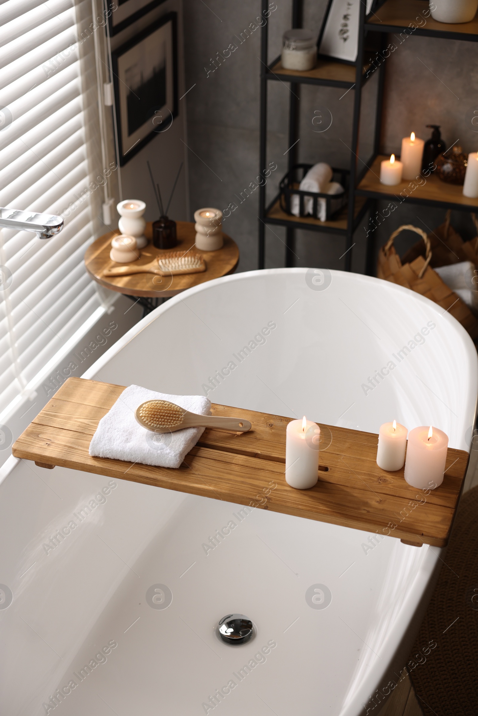 Photo of Wooden tray with burning candles, towel and brush on bathtub in bathroom