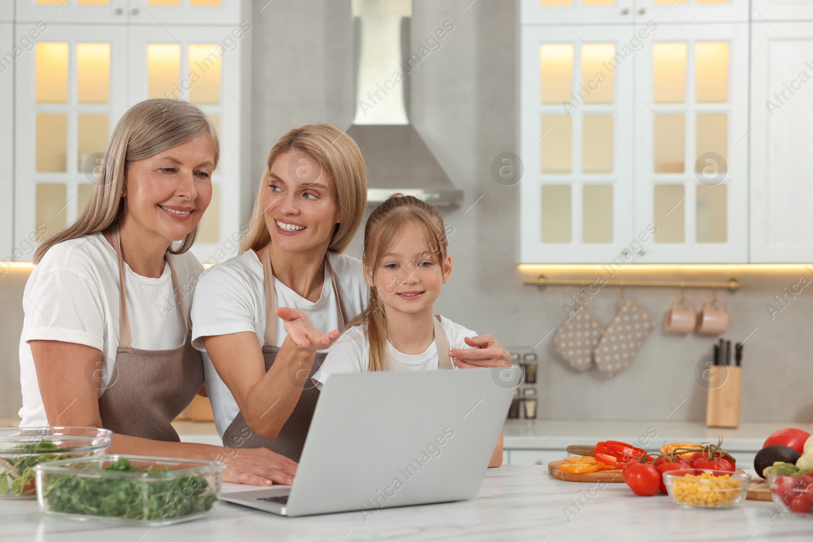 Photo of Three generations. Happy grandmother, her daughter and granddaughter using laptop in kitchen