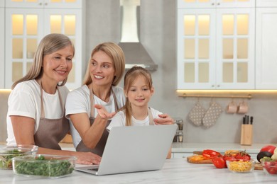 Photo of Three generations. Happy grandmother, her daughter and granddaughter using laptop in kitchen