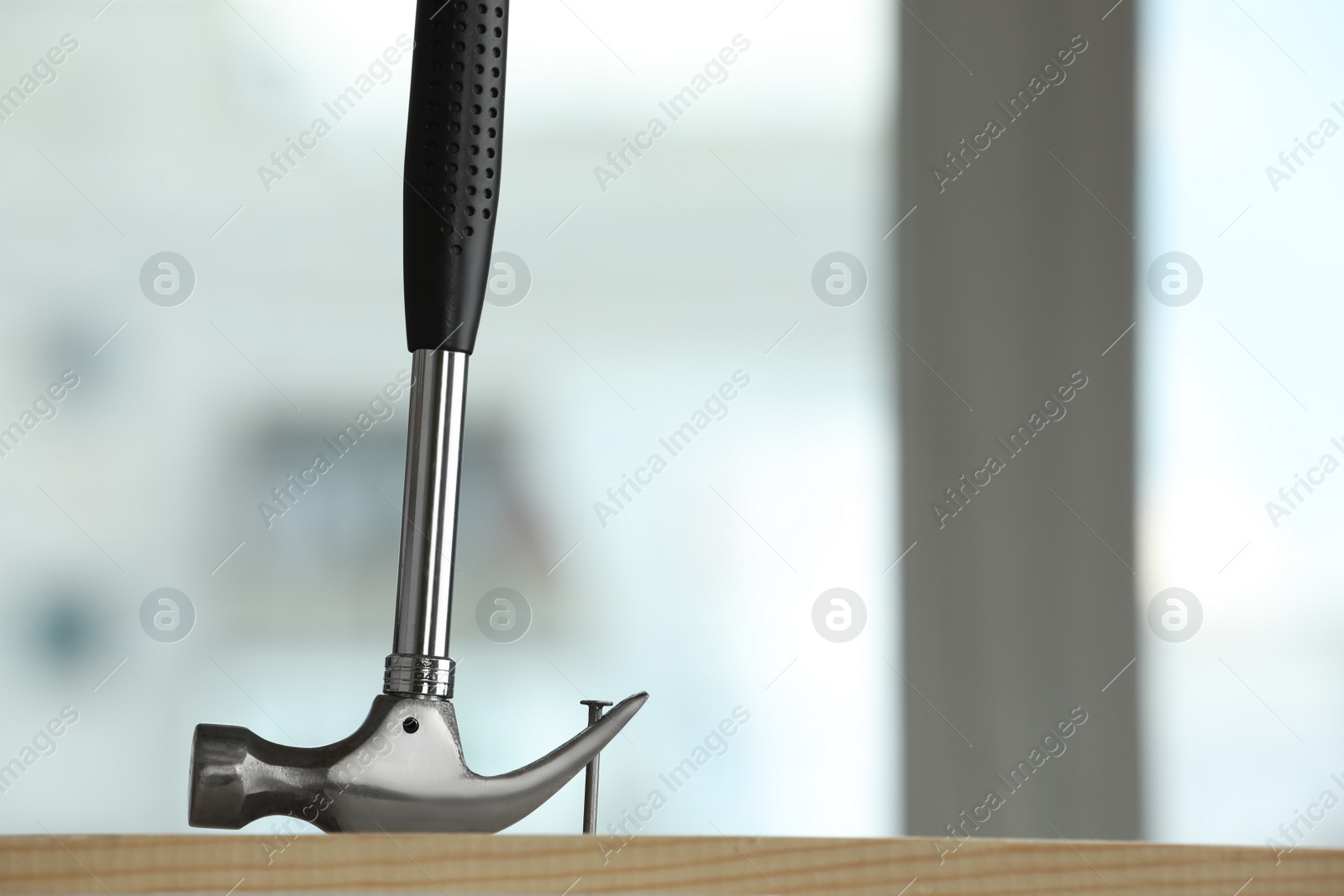 Photo of Hammer pulling metal nail out of wooden surface against blurred background indoors, space for text