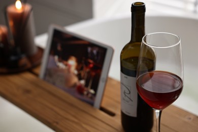 Photo of Wooden tray with tablet, glass of wine and bottle on bathtub in bathroom, selective focus