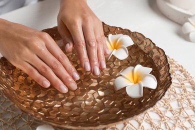 Photo of Woman soaking her hands in bowl with water and flowers on table, closeup. Spa treatment