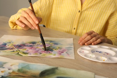 Photo of Woman painting flowers with watercolor at white wooden table, closeup. Creative artwork