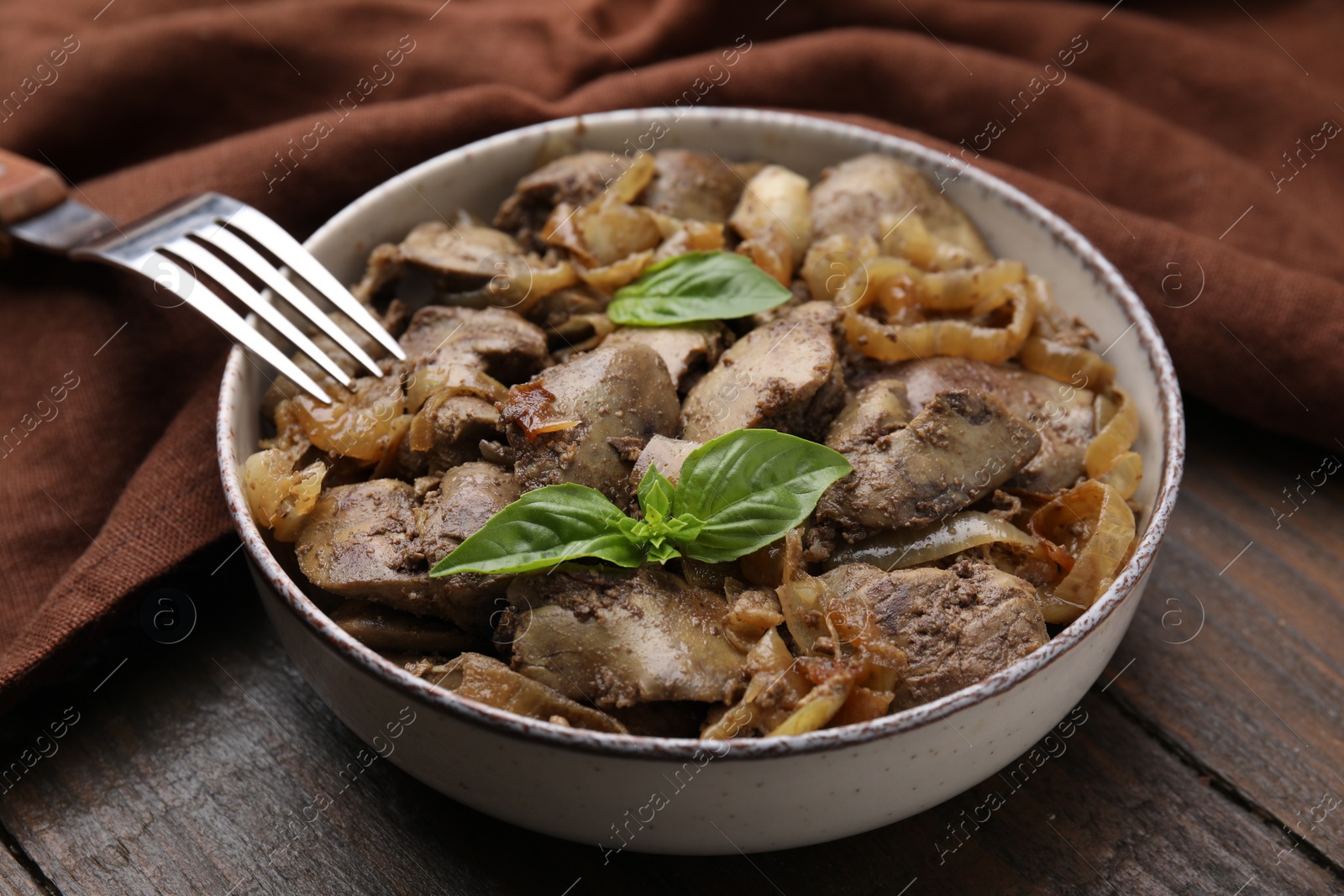 Photo of Delicious fried chicken liver with onion and basil in bowl on wooden table, closeup