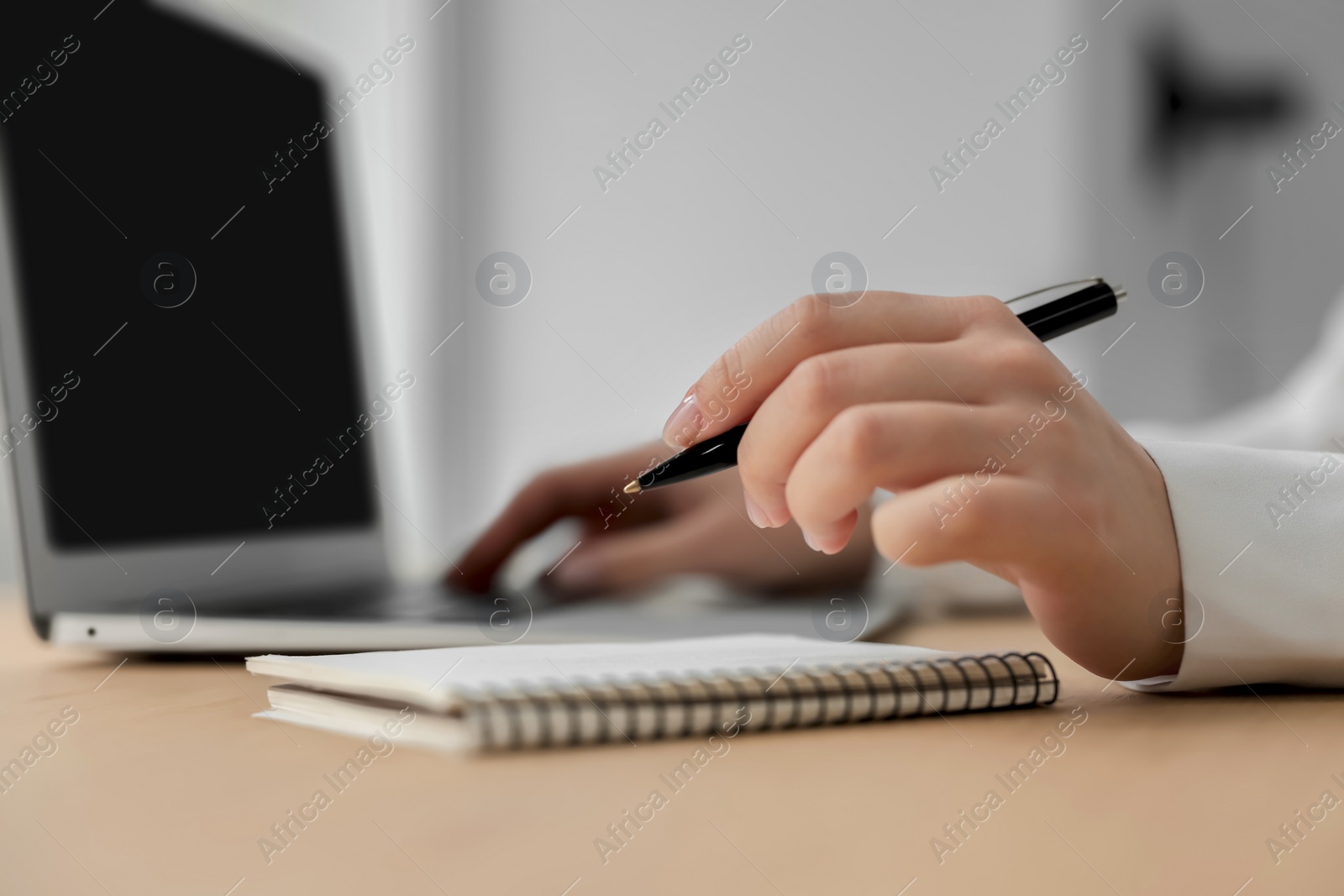 Photo of Woman with notebook and pen working on laptop at wooden table, closeup. Electronic document management