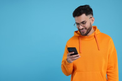 Happy young man using smartphone on light blue background, space for text