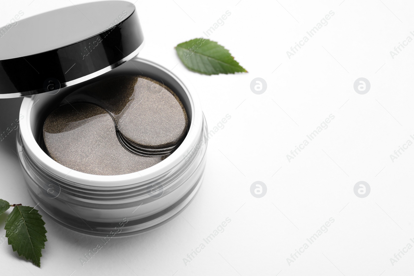Photo of Under eye patches in jar and green leaves on white background, space for text. Cosmetic product