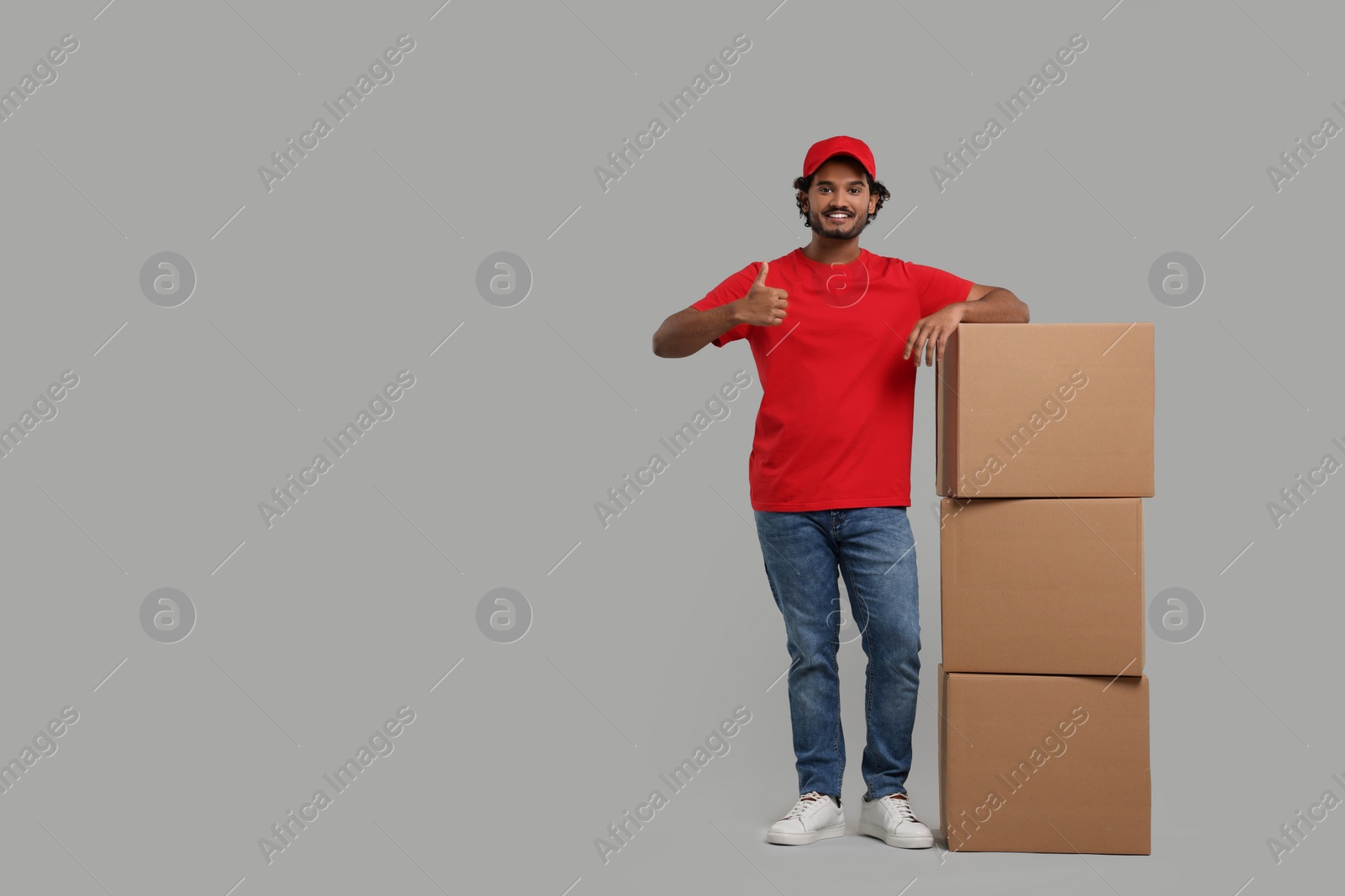 Photo of Happy courier with stack of parcels showing thumb up on grey background, space for text