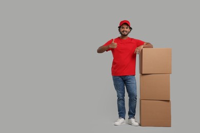 Photo of Happy courier with stack of parcels showing thumb up on grey background, space for text