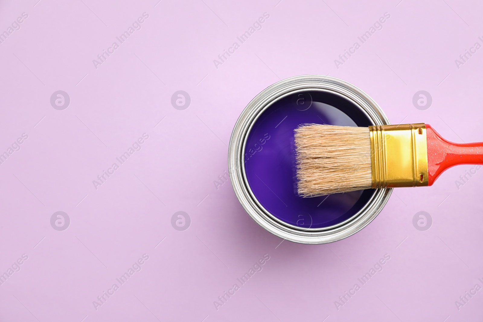 Photo of Open can with blue paint and brush on lilac background, top view. Space for text