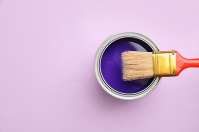 Open can with blue paint and brush on lilac background, top view. Space for text