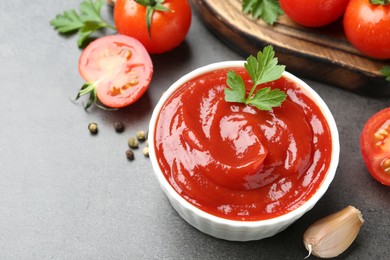 Photo of Delicious ketchup in bowl, tomatoes, parsley and garlic on grey table, closeup