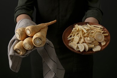 Woman holding whole and cut parsnips on black background, closeup