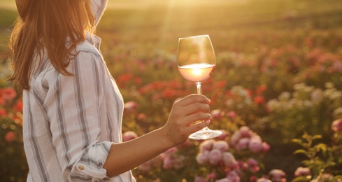 Woman with glass of wine in rose garden on sunny day, closeup. Space for text