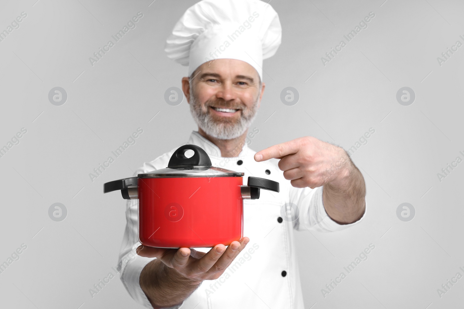 Photo of Happy chef in uniform pointing at cooking pot on grey background, selective focus