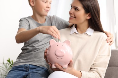 Photo of Boy with his mother putting money into piggy bank at home, closeup