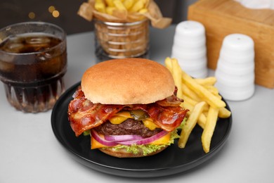 Tasty burger with bacon, vegetables and patty served with french fries and glass of refreshing drink on light grey table