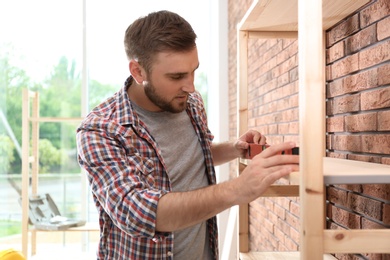 Photo of Young man working with construction level near wooden shelving unit indoors